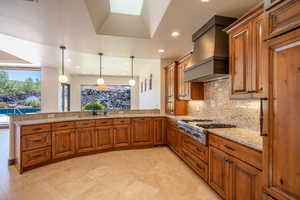 Kitchen with stainless steel gas stovetop, custom exhaust hood, hanging light fixtures, decorative backsplash, and kitchen peninsula