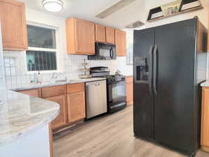 Kitchen featuring black appliances, sink, light wood-type flooring, tasteful backsplash, and light stone counters