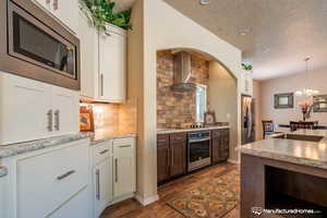 Kitchen with backsplash, wall chimney range hood, hanging light fixtures, appliances with stainless steel finishes, and white cabinetry