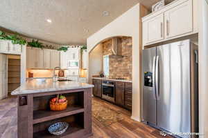 Kitchen with sink, wall chimney exhaust hood, dark wood-type flooring, a center island with sink, and appliances with stainless steel finishes