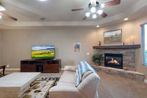 Living room with carpet flooring, ceiling fan, a stone fireplace, and a tray ceiling