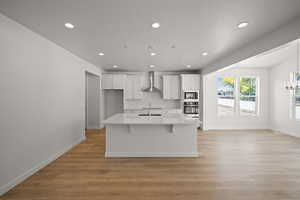 Kitchen with built in microwave, stainless steel oven, wall chimney range hood, a center island with sink, and white cabinets