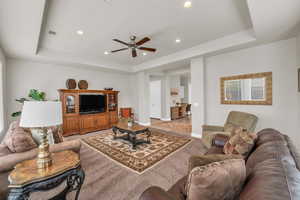 Carpeted living room with a raised ceiling, ceiling fan, and a healthy amount of sunlight