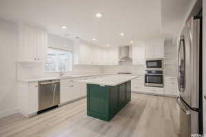 Kitchen with wall chimney exhaust hood, a kitchen island, appliances with stainless steel finishes, white cabinets, and light wood-type flooring