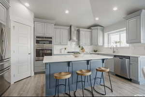 Kitchen with gray cabinetry, lofted ceiling, wall chimney range hood, appliances with stainless steel finishes, and a kitchen island