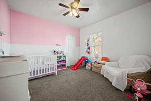 Bedroom featuring dark colored carpet, ceiling fan, a crib, and a textured ceiling