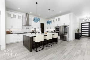 Kitchen featuring appliances with stainless steel finishes, wall chimney exhaust hood, decorative light fixtures, white cabinetry, and an island with sink