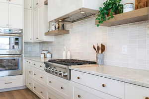 Kitchen with light stone countertops, white cabinetry, tasteful backsplash, premium range hood, and appliances with stainless steel finishes