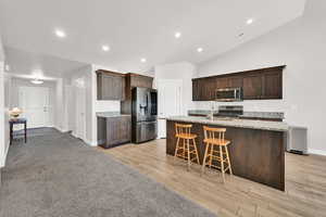 Kitchen with light stone countertops, stainless steel appliances, a center island with sink, light hardwood / wood-style floors, and lofted ceiling