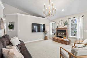 Living room with lofted ceiling, an inviting chandelier, a brick fireplace, ornamental molding, and carpet floors