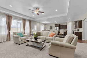 Living room with a tray ceiling, light colored carpet, and ceiling fan with notable chandelier