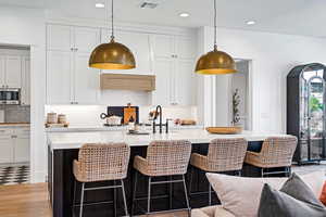 Kitchen featuring stainless steel microwave, a center island with sink, white cabinets, and decorative light fixtures