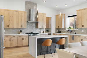 Kitchen featuring wall chimney exhaust hood, quarts counter tops, waterfall edge, vaulted ceiling, decorative backsplash, and light brown cabinetry