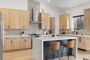 Kitchen featuring wall chimney exhaust hood, quarts counter tops, waterfall edge island, vaulted ceiling, decorative backsplash, and light brown cabinetry