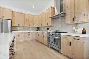 Kitchen featuring wall chimney exhaust hood, quarts counter tops, waterfall edge island, vaulted ceiling, decorative backsplash, and light brown cabinetry