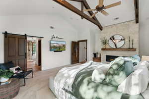 Bedroom with ceiling fan, a barn door, beamed ceiling, a large fireplace, and light colored carpet