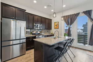 Kitchen with sink, light stone counters, an island with sink, pendant lighting, and appliances with stainless steel finishes