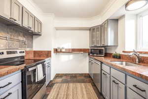 Kitchen with gray cabinetry, crown molding, sink, light stone countertops, and stainless steel appliances