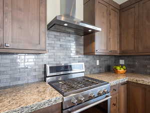 Kitchen featuring wall chimney exhaust hood, light stone countertops, backsplash, and stainless steel range