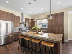 Kitchen featuring a center island, wall chimney range hood, hanging light fixtures, light stone countertops, and stainless steel appliances