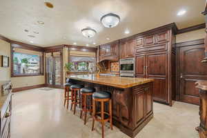 Kitchen featuring a kitchen breakfast bar, light stone counters, stainless steel oven, a kitchen island with sink, and crown molding