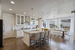 Kitchen with wall chimney exhaust hood, stainless steel appliances, decorative backsplash, a center island with sink, and white cabinets