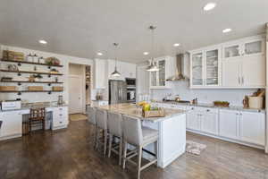 Kitchen with a center island, wall chimney exhaust hood, stainless steel appliances, light stone counters, and white cabinets