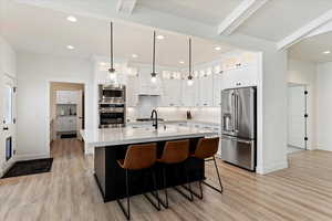 Kitchen with a kitchen island with sink, beamed ceiling, decorative light fixtures, white cabinetry, and stainless steel appliances