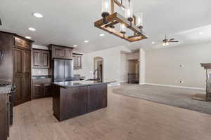 Kitchen featuring light wood-type flooring, dark brown cabinets, stainless steel appliances, dark stone countertops, and an island with sink