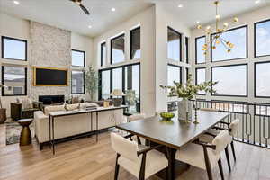 Dining room with light wood-type flooring, a towering ceiling, a stone fireplace, and ceiling fan with notable chandelier