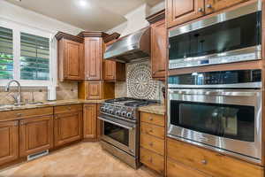 Kitchen featuring backsplash, exhaust hood, sink, light stone countertops, and appliances with stainless steel finishes