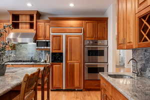 Kitchen featuring sink, built in appliances, decorative backsplash, wall chimney exhaust hood, and light stone countertops