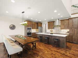 Kitchen with wall chimney exhaust hood, stainless steel appliances, a kitchen breakfast bar, pendant lighting, and a center island with sink