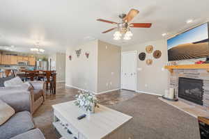 Carpeted living room featuring a stone fireplace and ceiling fan with notable chandelier