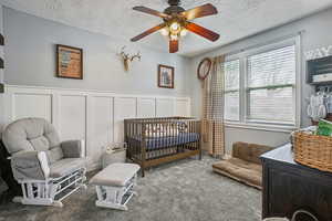 Bedroom with ceiling fan, carpet floors, a textured ceiling, multiple windows, and a crib