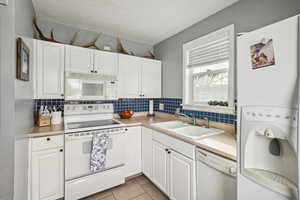 Kitchen featuring backsplash, white appliances, sink, light tile patterned floors, and white cabinetry