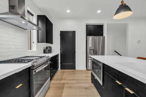 Kitchen with light wood-type flooring, backsplash, wall chimney exhaust hood, stainless steel appliances, and pendant lighting