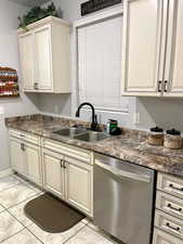 Kitchen featuring cream cabinets, sink, stainless steel dishwasher, dark stone countertops, and light tile patterned flooring