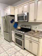 Kitchen featuring appliances with stainless steel finishes, light tile patterned floors, dark stone counters, and cream cabinets