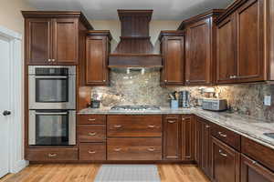 Kitchen with premium range hood, light stone countertops, light hardwood / wood-style floors, and appliances with stainless steel finishes