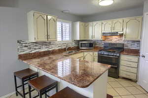 Kitchen featuring black gas range, dark stone counters, sink, light tile patterned flooring, and kitchen peninsula