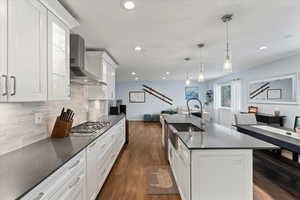 Kitchen with dark wood-type flooring, wall chimney range hood, pendant lighting, a center island with sink, and white cabinets