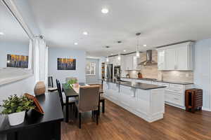 Kitchen featuring wall chimney exhaust hood, an island with sink, pendant lighting, white cabinets, and appliances with stainless steel finishes