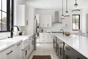 Kitchen featuring light stone counters, white cabinetry, decorative light fixtures, and wall chimney range hood