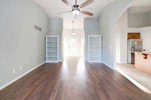 Unfurnished living room with high vaulted ceiling, arched walkways, a ceiling fan, dark wood-style floors, and baseboards