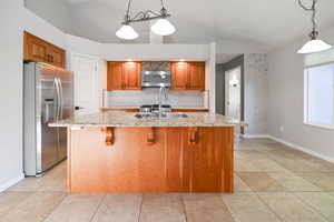 Kitchen with stainless steel fridge with ice dispenser, decorative backsplash, light stone countertops, brown cabinetry, and baseboards