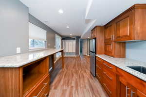 Kitchen with freestanding refrigerator, brown cabinetry, light wood-style flooring, light stone countertops, and recessed lighting