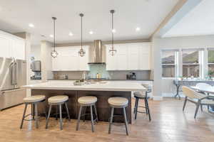 Kitchen with hanging light fixtures, a center island with sink, high end fridge, and wall chimney range hood