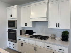 Kitchen with stainless steel appliances, tasteful backsplash, light stone counters, white cabinets, and custom exhaust hood