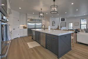 Kitchen with appliances with stainless steel finishes, light wood-type flooring, a kitchen island with sink, white cabinets, and hanging light fixtures